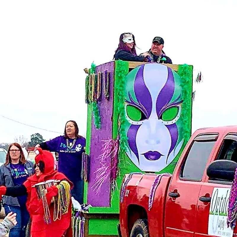 Mardi Gras float with large mask decoration, people throwing beads from the float and truck.