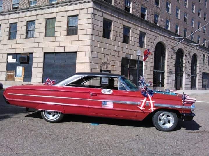Red classic car with American flag decorations parked on a street in front of a stone building.