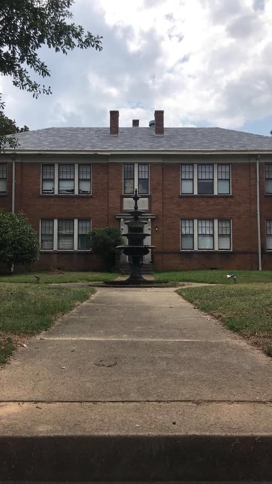 Brick apartment building with fountain in front, sidewalk leading up to the entrance, cloudy sky.