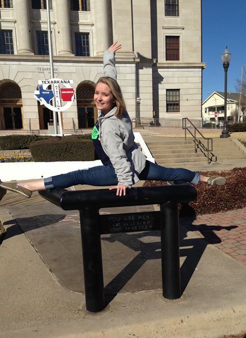 Woman in grey sweatshirt and jeans doing the splits on a black bench outside a building.