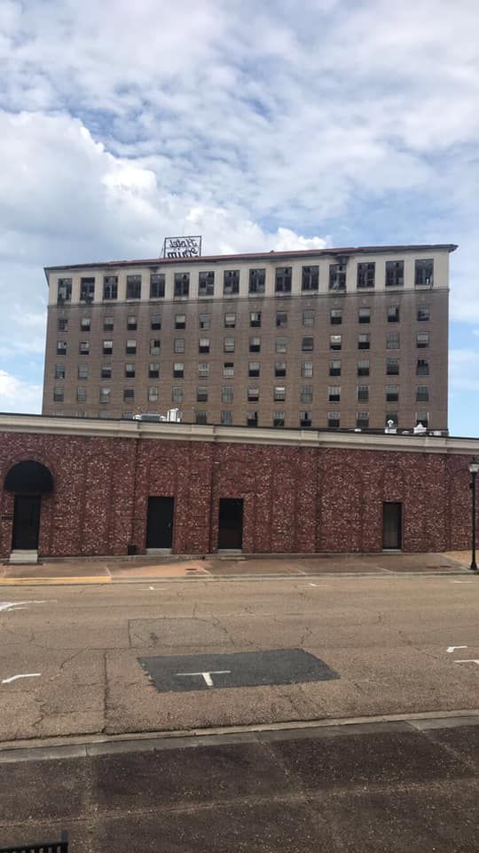 Seven-story brick building with many windows, topped with a sign. Below is a one-story brick building. Overcast sky.