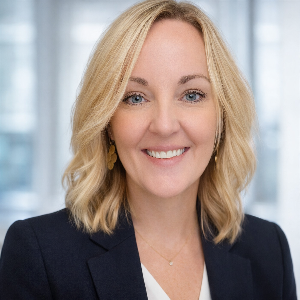 Woman with blonde hair, smiling at the camera, wearing a blazer, in a bright office setting.