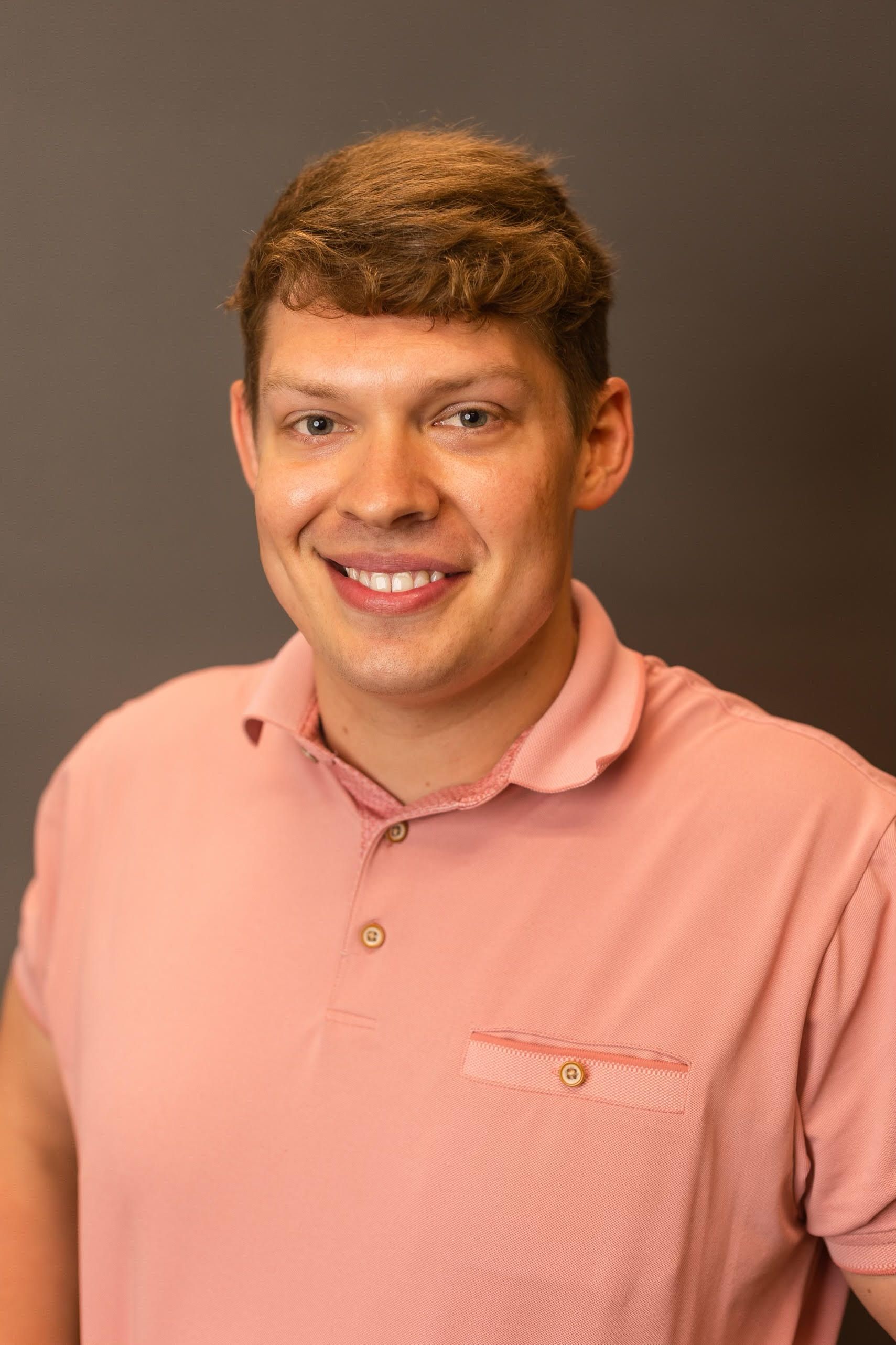 Man in a pink polo shirt smiling at the camera against a grey backdrop.