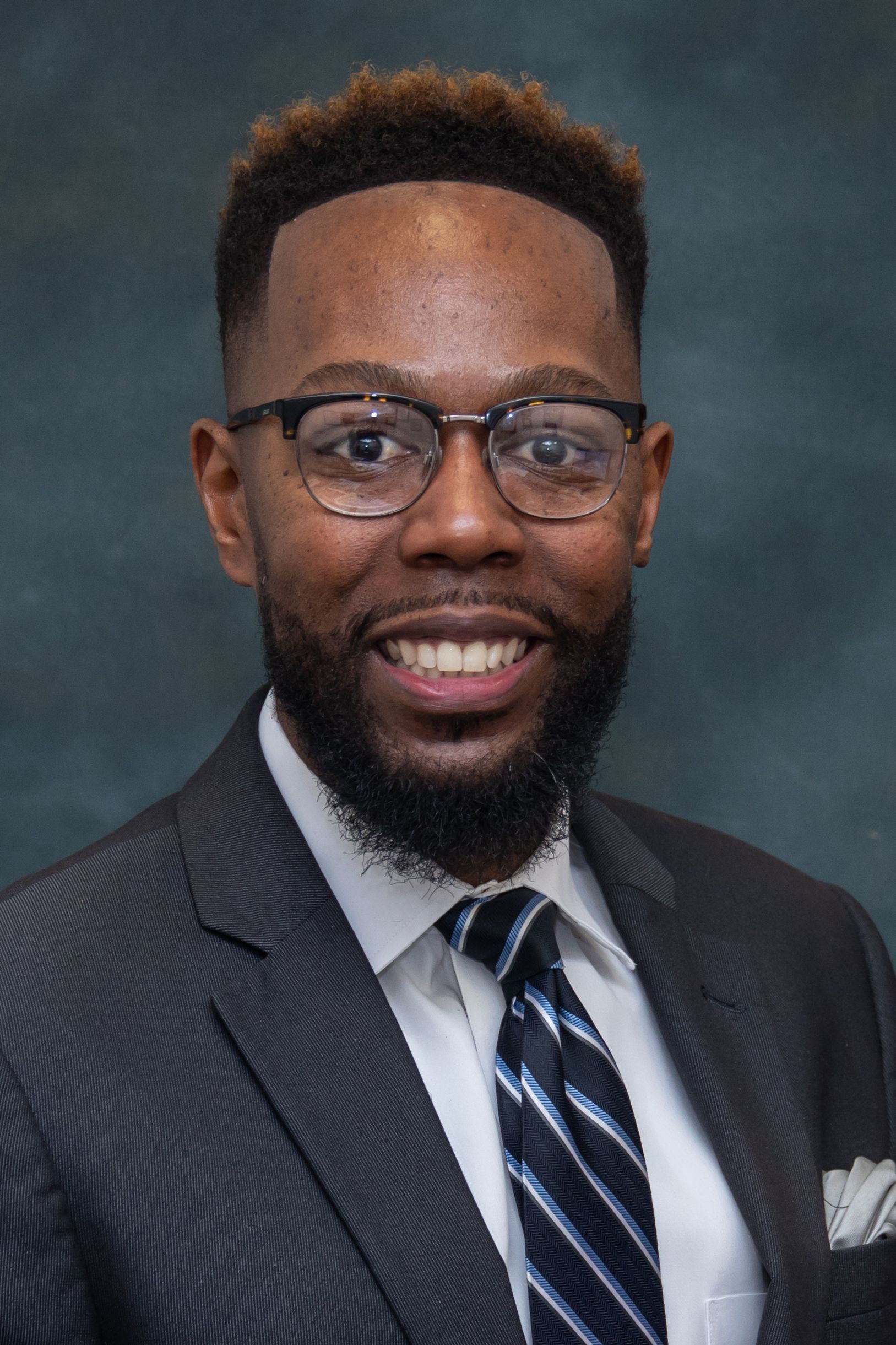 Man wearing glasses, suit, and tie smiling at the camera.