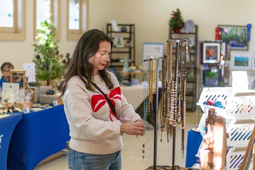Woman browsing necklaces at an indoor craft fair; she's wearing a sweater with a bow design.