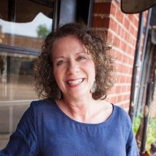 Woman with curly brown hair smiling outdoors, wearing a blue shirt.