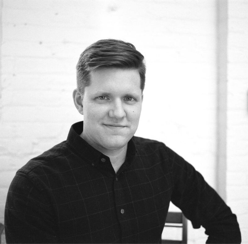 Man in a black shirt smiles, seated against a white brick wall.