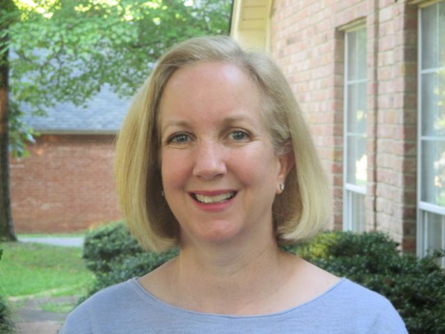 Woman with blonde hair smiling outdoors in front of a brick building and greenery.