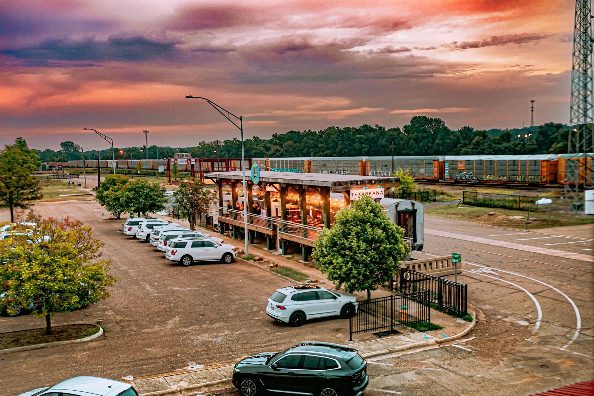 A restaurant with outdoor seating and a parking lot at sunset, near a railroad.