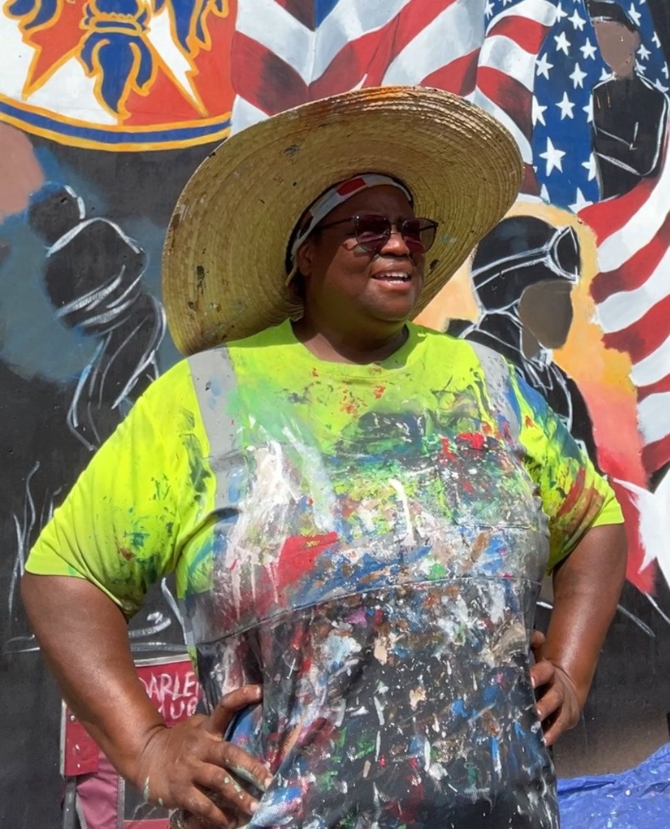 Woman in paint-splattered shirt and straw hat stands before a mural, smiling, hands on hips.