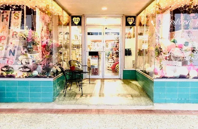 Storefront with glass windows, teal tiled base, and a small table. Displays of colorful items inside.