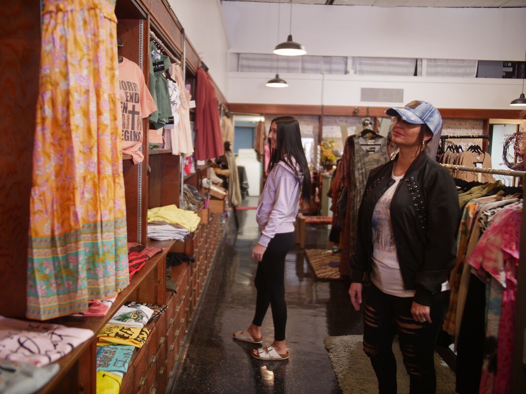 Two women browsing clothing racks in a boutique. Shelves filled with apparel line the wall.