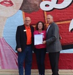 Three people stand in front of a mural, a woman holding a document, being given by a man, the other man standing to the left.