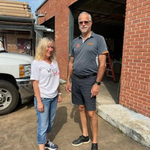 Woman and man standing by a brick building and a truck; man wearing shorts and a polo, woman in jeans and a t-shirt.