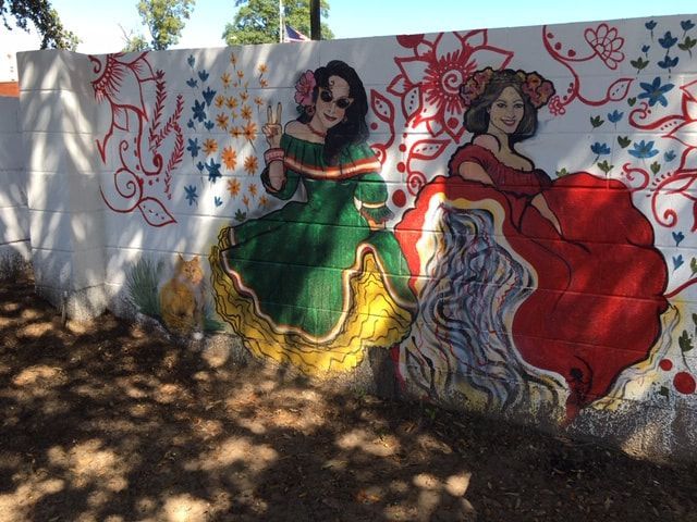 Mural of two women in vibrant dresses, surrounded by floral designs. Green and red dresses. Sunlight on wall.