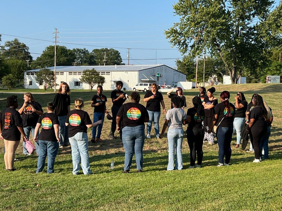 Group of people in a circle outdoors, some wearing shirts with a rainbow graphic, near a building on a grassy field.