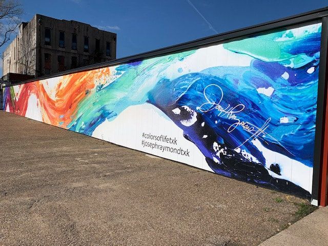 Colorful abstract mural on a long wall with text and a signature; building in background.