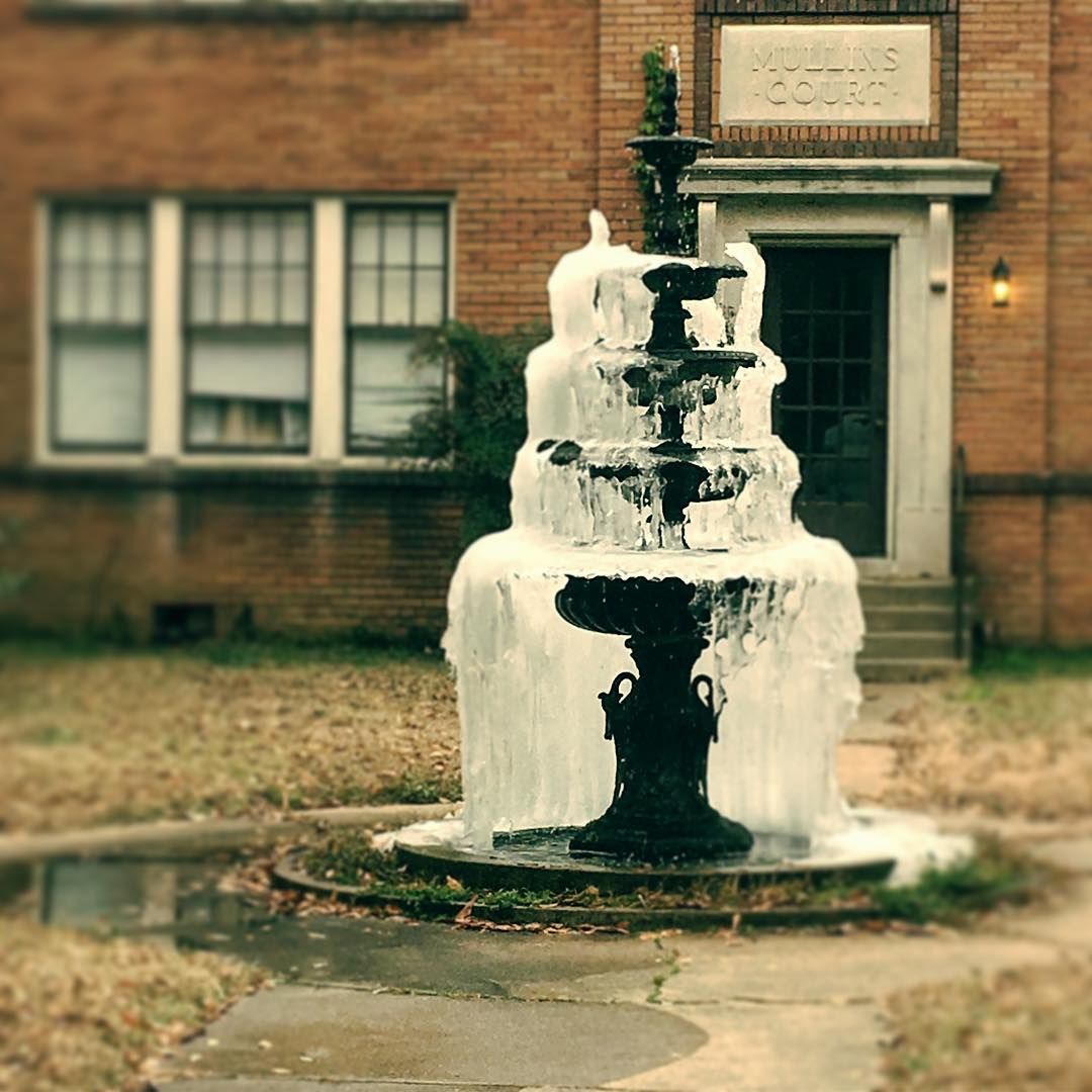 Frozen tiered fountain in front of a brick building. Ice covers the structure, with a cloudy sky in the background.