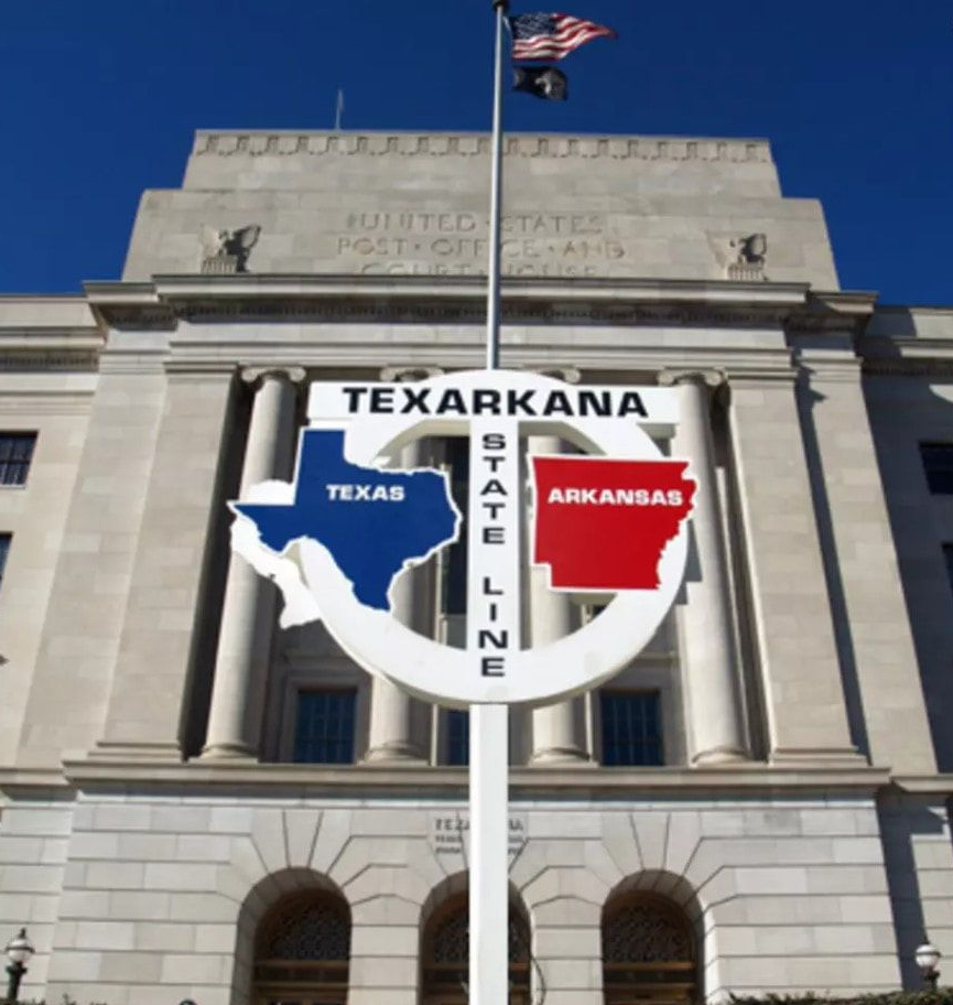 Texarkana state line sign with Texas (blue) and Arkansas (red) outlines, in front of the US Post Office building under a blue sky.
