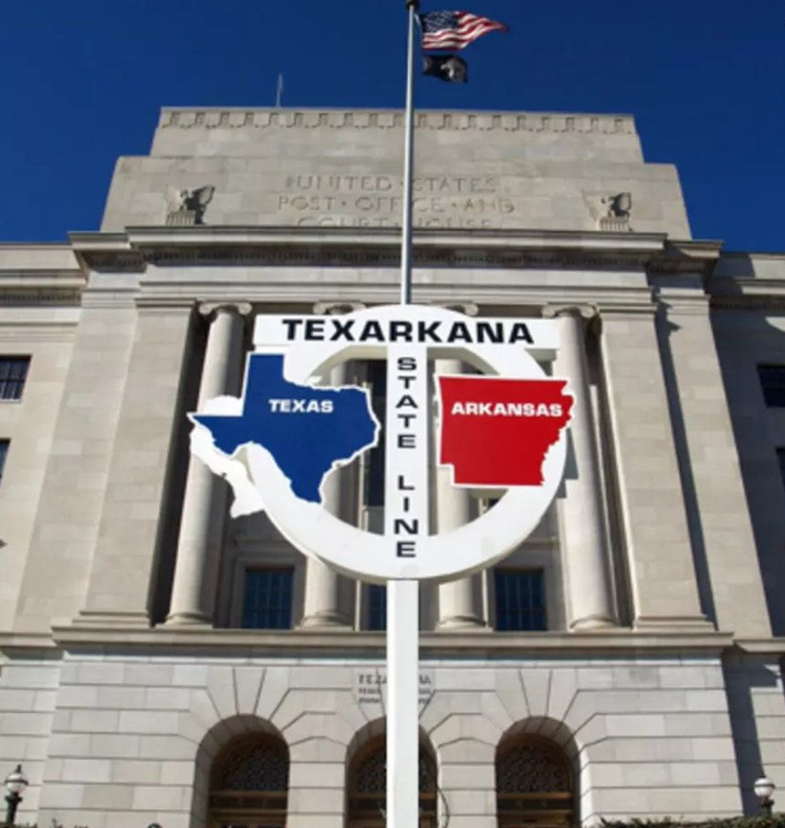 Texarkana state line sign with Texas and Arkansas outlines, in front of a post office building.