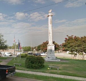 Monument with eagle on top in a grassy area with flags, benches, and parked vehicles.
