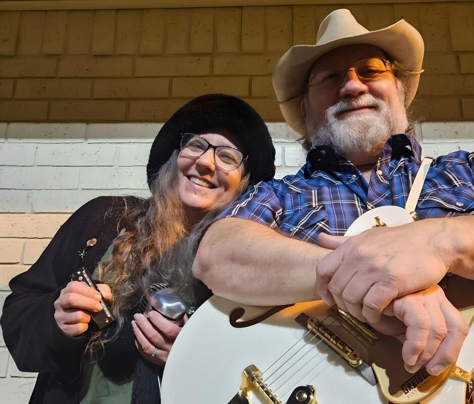 Two musicians smiling. Woman holds harmonicas, man holds a white guitar, both wear hats, outside at night.