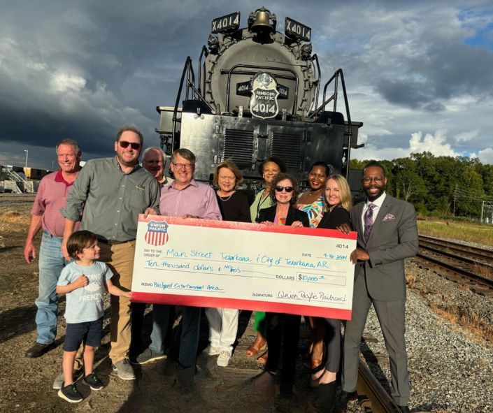 People holding a large check in front of a train, near train tracks. Cloudy sky in the background.