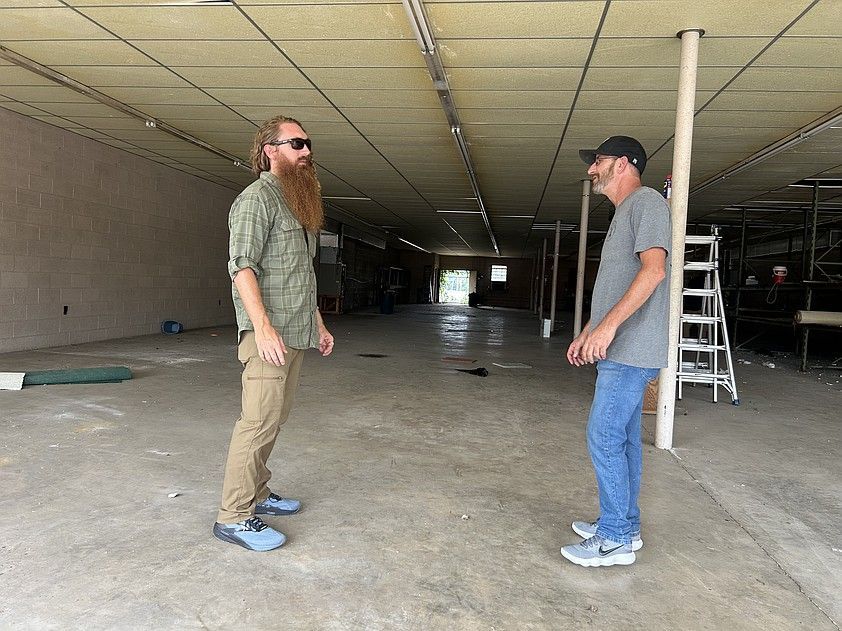 Two men facing each other in an empty, dilapidated building. One with a long beard, the other wearing a baseball cap.