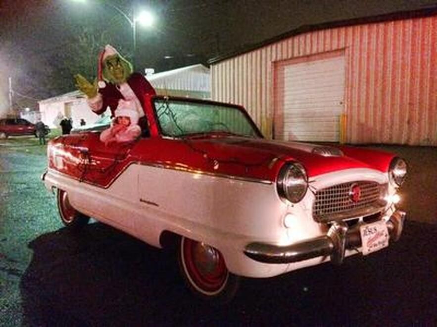 Grinch in Santa suit waves from a red and white vintage convertible, evening setting.
