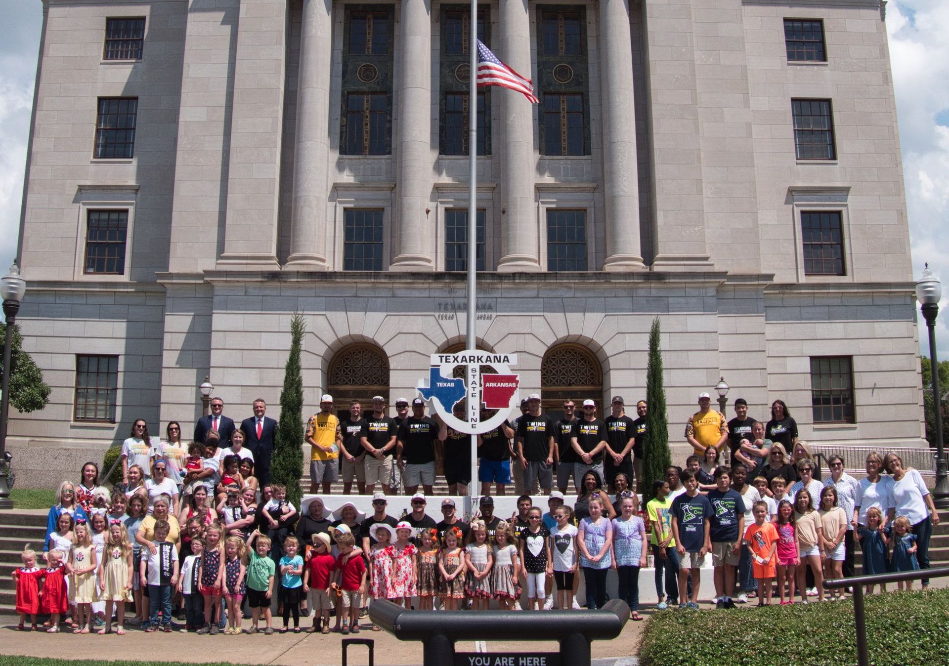 Large group of people posing in front of a light-colored building with a flagpole and an American flag.