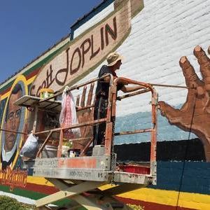 Artist on lift painting a mural of Scott Joplin and a hand. Bright colors, exterior setting.