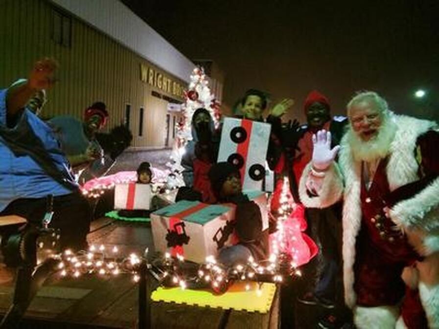 Santa and children on a holiday float, waving, in front of a building with lights.