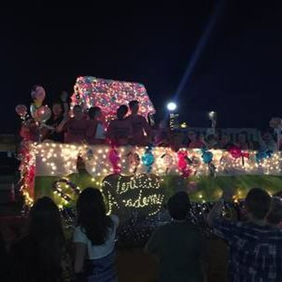 People on a brightly lit float at night, possibly a parade. The float says