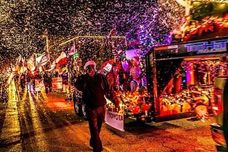 Nighttime Christmas parade with illuminated floats and falling snow, a person walks near a Toyota sign.