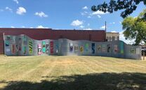 Long, gray, wavy outdoor wall featuring colorful painted panels with text and images, in a grassy area under a blue sky.