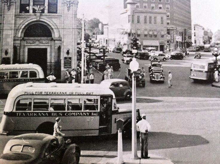 Vintage street scene with buses, cars, and pedestrians at an intersection. Bank building, storefronts, and a tall building are visible.