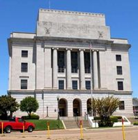 Large, light-colored building with columns, steps, and windows; a red vehicle is parked in front.