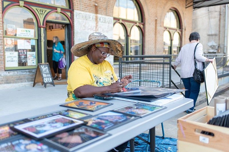 Woman at outdoor table selling framed art, wearing a hat, yellow shirt. Building background.