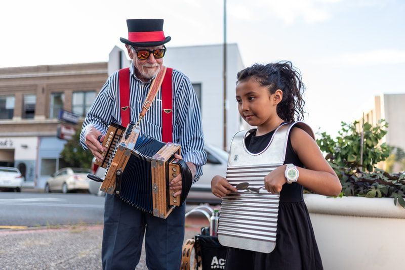 Man in top hat playing accordion, girl playing washboard outside; brick building in background.