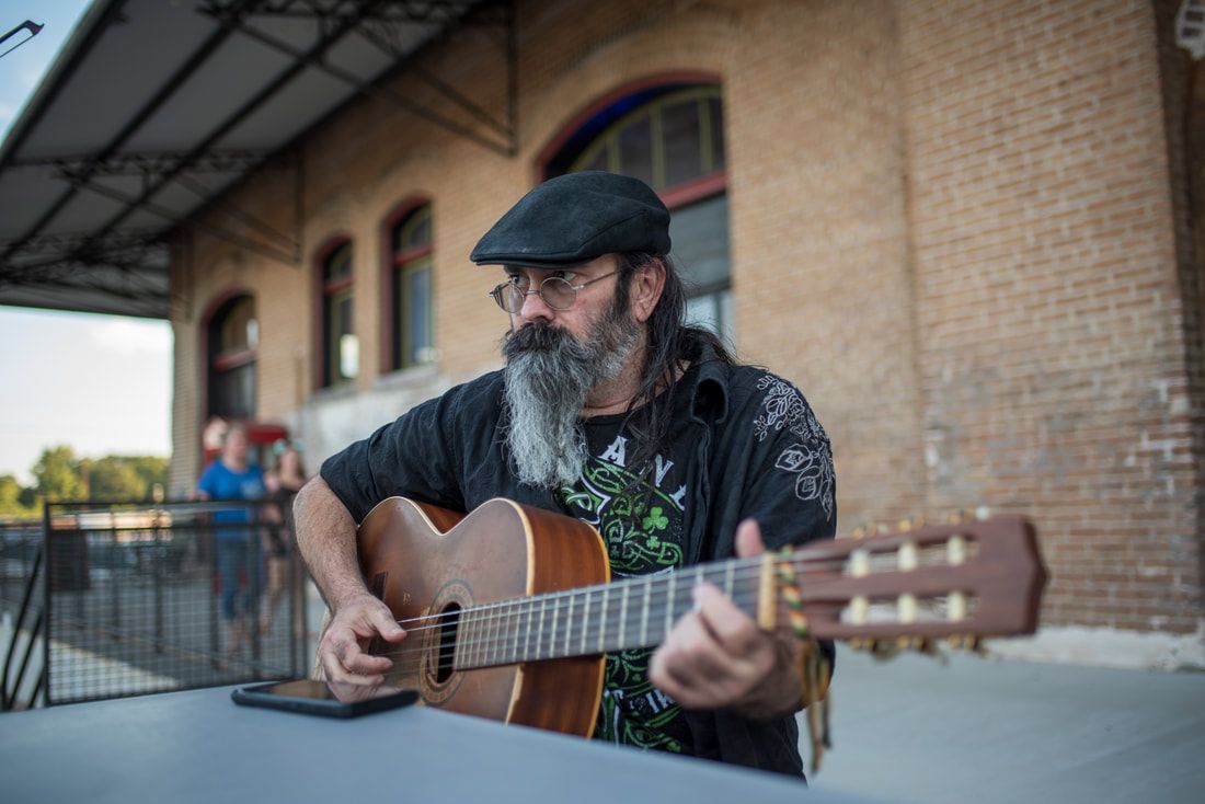 Man with a graying beard playing an acoustic guitar outdoors. Brick building in the background.