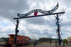 Sign for Front Street Festival Plaza, with artistic metalwork, cloudy sky, and a train car.