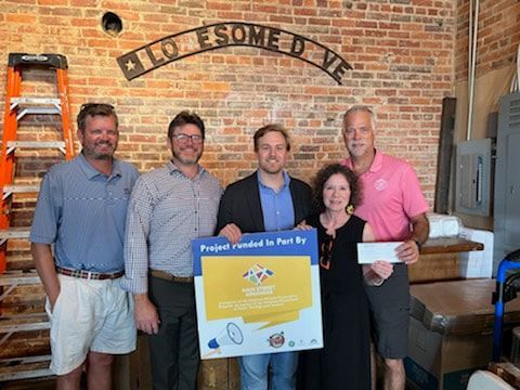Group of five people holding a check and a sign; brick wall in background.
