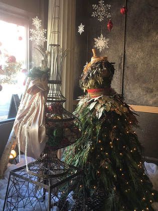 Christmas-themed window display with a mannequin dressed as a tree, alongside a decorative iron birdcage. Snowflakes and lights.