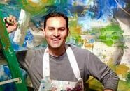 Man in grey shirt and apron smiles in front of colorful abstract painting.