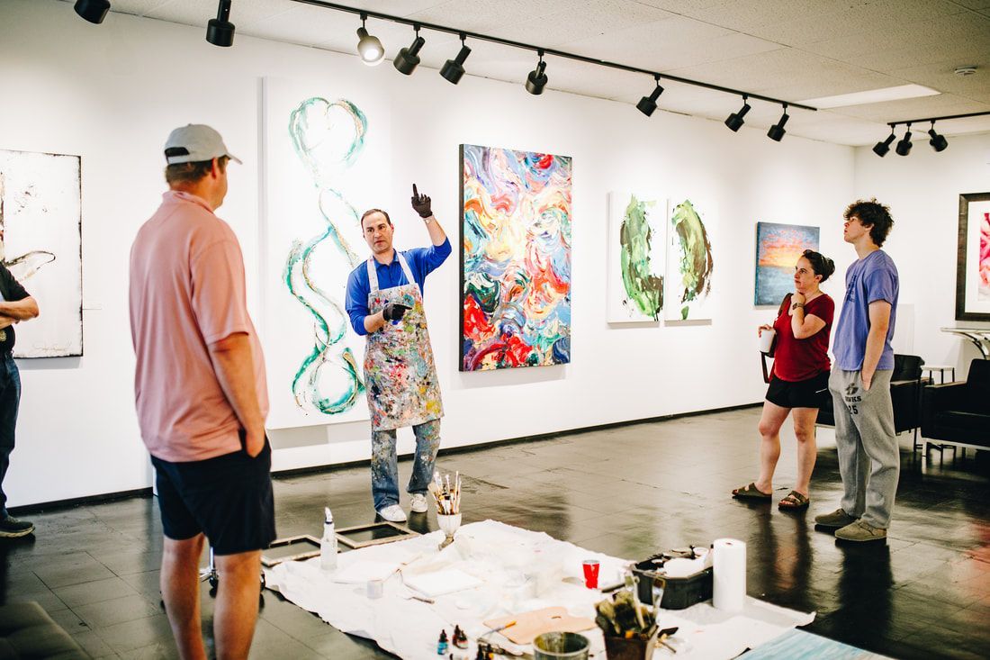 Man with apron points at painting in art gallery, explaining to visitors. Paintings and art supplies visible.
