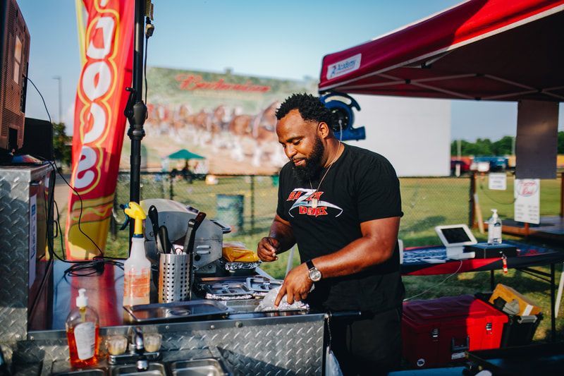 Man preparing food at a food stand with a red canopy, Budweiser mural backdrop.