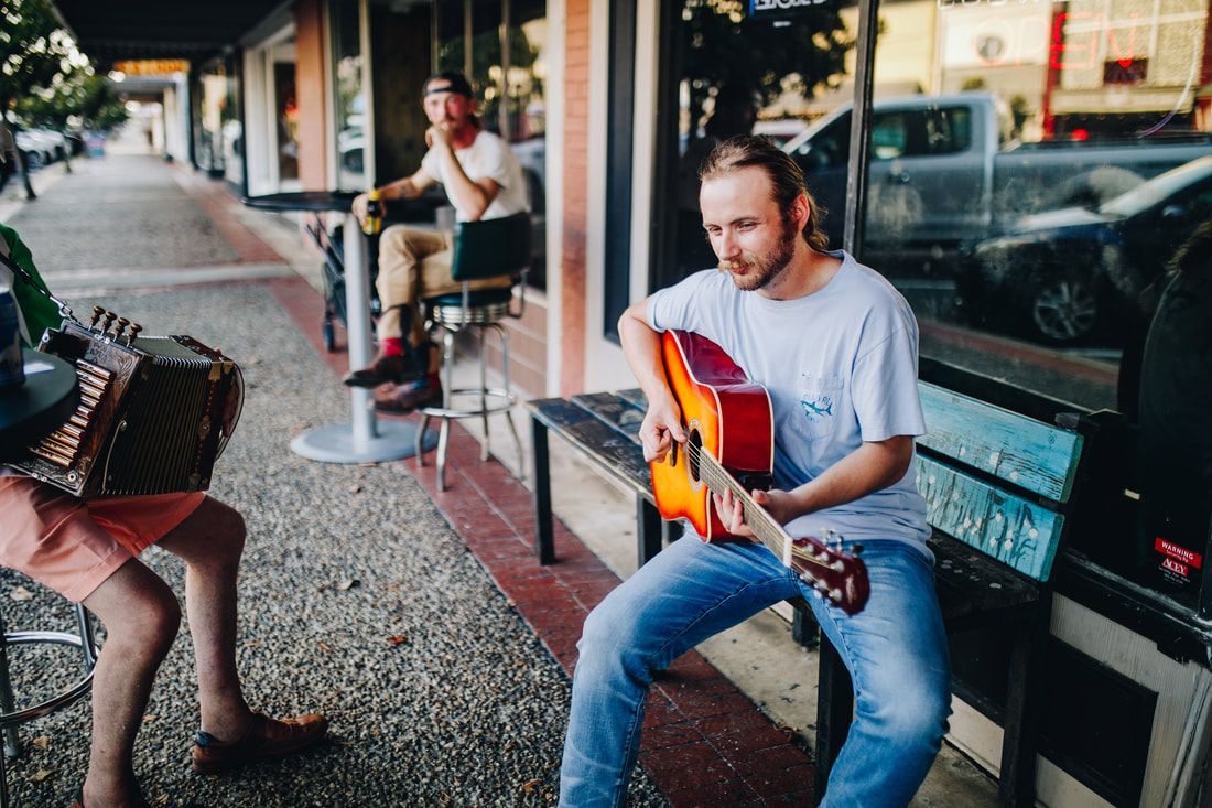 Street musicians: guitarist, accordion player, and observer seated outdoors near storefront.