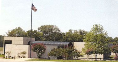 A building with a US flag. Tan walls, arched roof, and trees in the background.