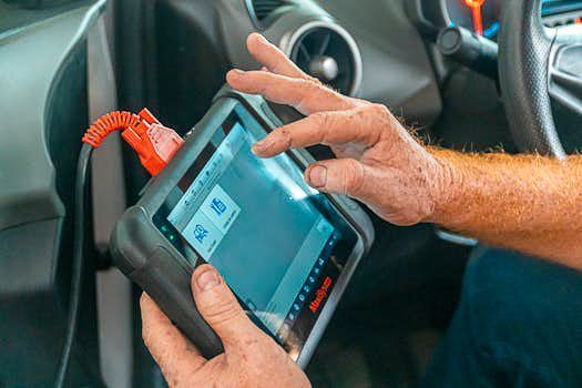 Mechanic at S&S Automotive Service using a diagnostic scanner in a car. The device is connected; showing a screen with icons.
