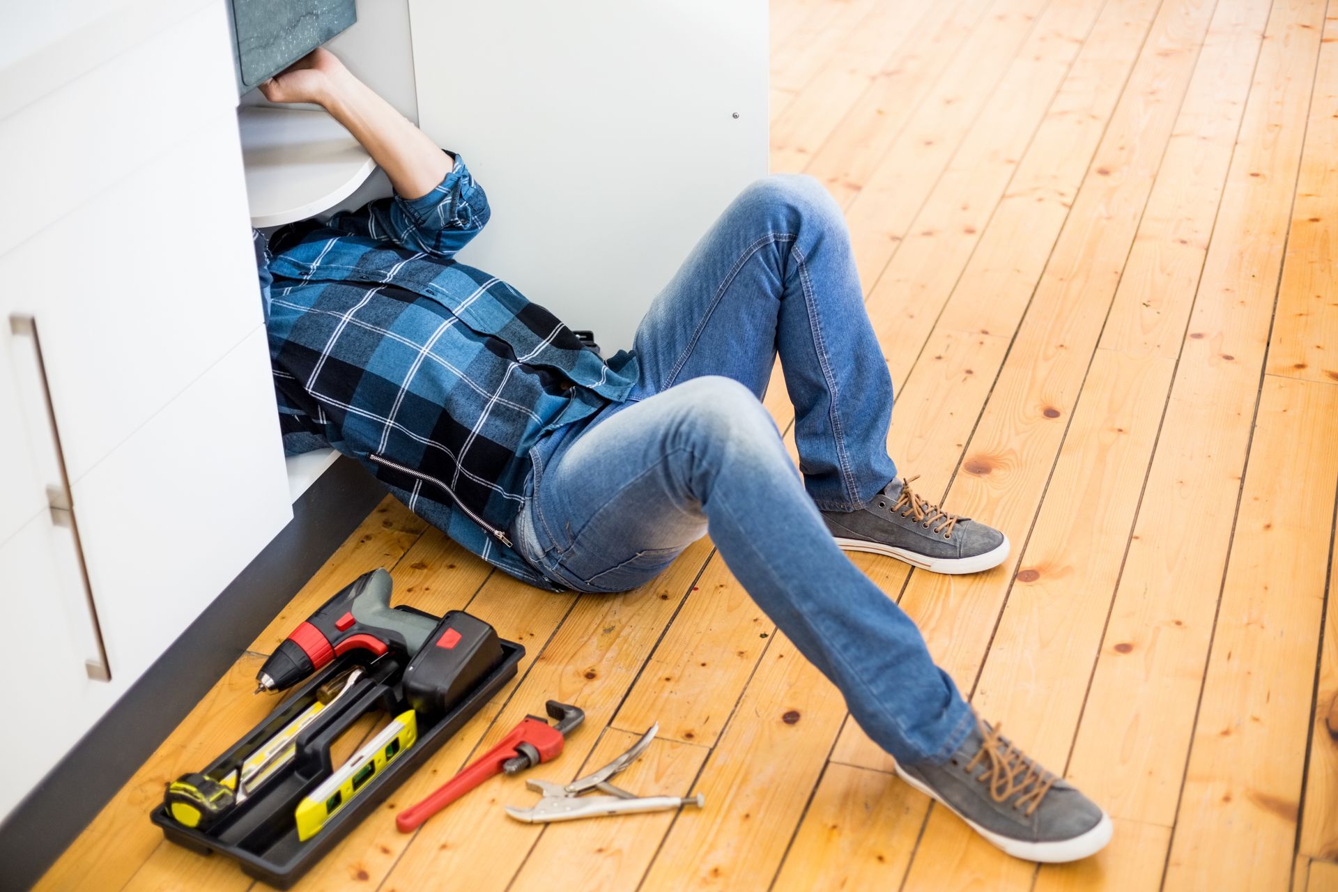 Person lying on floor fixing plumbing under a wooden cabinet with a tool bag nearby.